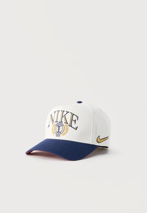 Witte en marineblauwe Nike pet met geborduurd gouden logo, tekst "NIKE 1972" en blauwe gekromde klep op een witte achtergrond.