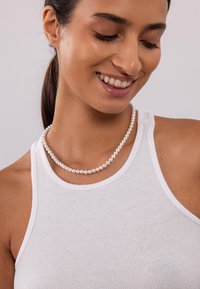Parelketting met uniforme ronde witte parels, gedragen over een witte geribbelde tanktop. Glad oppervlak en subtiele glans.