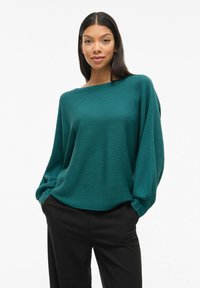 Pull en maille côtelée bleu sarcelle avec un large col et des épaules tombantes, associé à un pantalon noir. Texture douce et coupe décontractée. Pas de motifs ni d'accents.