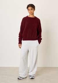 Jeune femme debout, portant un pull pelucheux bordeaux, un pantalon large gris clair et des baskets grises, sur un fond clair uni.