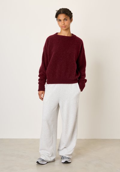 Jeune femme debout, portant un pull pelucheux bordeaux, un pantalon large gris clair et des baskets grises, sur un fond clair uni.