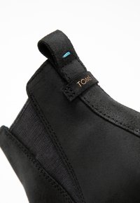 Bottine en suédine noire avec des panneaux élastiques sur les côtés, une finition texturée, des coutures détaillées et une petite touche de bleu sur la languette.