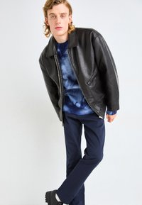 Veste en cuir noir avec un col, portée sur un pull bleu tie-dye, associée à un pantalon marine et des chaussures noires.