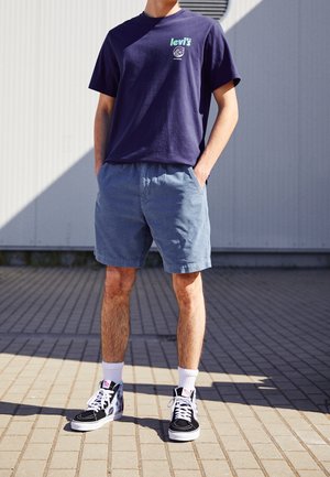 Mand stående på brolagt underlag iført navy Levi's t-shirt, lyseblå shorts, hvide sokker og sorte høje sneakers med hvide snørebånd.