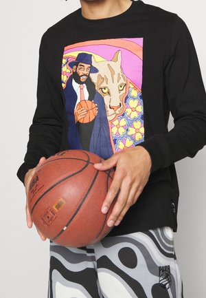 Person hält einen Basketball und trägt ein schwarzes Shirt mit einer bunten Grafik eines Mannes mit Hut, der einen Basketball neben einer großen Katze und einem floralen Hintergrund hält.