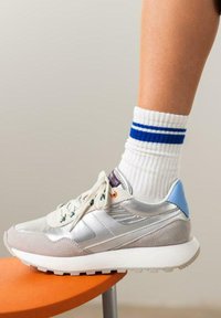 Zapatillas grises y plateadas con material de ante y brillante, acento azul en el talón. Calcetines blancos con rayas azules, pie apoyado en un taburete naranja.