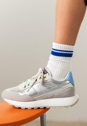 Baskets grises et argentées avec des matériaux en daim et scintillants, accent bleu au niveau du talon. Chaussettes blanches côtelées avec des rayures bleues, pied reposant sur un tabouret orange.