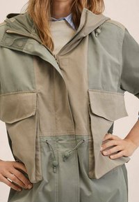 Veste légère bicolore en vert pâle et beige. Dotée d'une capuche ajustable, de plusieurs poches avant et d'un cordon de serrage à la taille pour un ajustement personnalisé.