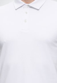Tricou polo alb cu trei nasturi și un logo „BOSS” subtil pe partea stângă a pieptului, purtat de o persoană.