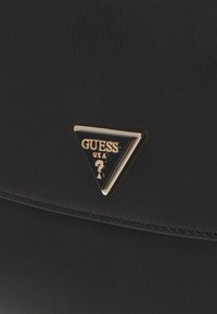 Borsa in finta pelle nera con piastra logo triangolare oro e nera con la scritta "GUESS". Superficie strutturata e dettagli di cucitura visibili.