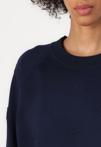 Mørkeblå hettegenser med teksturert mønster, med tonale logoaksenter. Ribbestrikket rund hals og raglanermer for en avslappet passform.