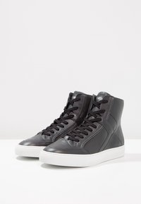 Baskets montantes en cuir noir lisse avec une semelle en caoutchouc blanche ; elles présentent un design à lacets et des accents de coutures sur les côtés.