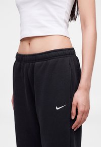 Γυναίκα που φοράει μαύρη φόρμα Nike και λευκό κοντό τοπ, εμφανιζόμενη από την κοιλιά μέχρι τα μέσα του μηρού σε απλό φόντο.