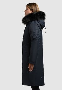 Cappotto smanicato lungo di un blu navy scuro con cappuccio in pelliccia sintetica, tasche laterali e un patch testurizzato sul braccio. Vestibilità dritta, tessuto liscio e lunghezza caviglia.
