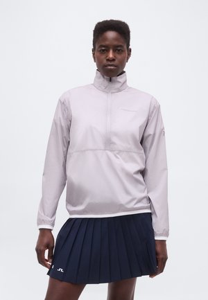 Γυναίκα που φοράει ανοιχτό γκρι μπουφάν με φερμουάρ μέχρι τη μέση και μια σκούρα μπλε πλισέ φούστα, στέκεται μπροστά σε απλό λευκό φόντο.