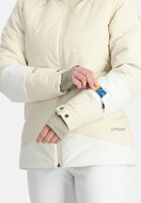 Beige, wattierte Jacke mit kontrastierenden weißen Einsätzen, enganliegenden Bündchen und einer Reißverschlusstasche mit einem blauen und gelben Etikett. Markenlogo sichtbar.