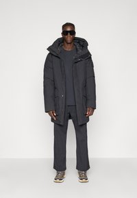 Black insulated parka with a hood, featuring a zip front, side pockets, and a relaxed fit paired with matching dark clothing and sneakers.