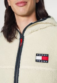 Sudadera de forro polar color crema con una cremallera azul marino, que presenta un logo de parche negro etiquetado "TOMMY JEANS" y detalles en rojo y blanco.
