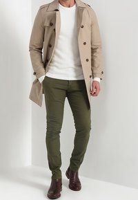 Beige Trenchcoat mit doppelter Knopfleiste, braunen Knöpfen und Klappentaschen. Kombiniert mit einem strukturierten weißen Pullover, olivgrünen Hosen und dunkelbraunen Stiefeln.