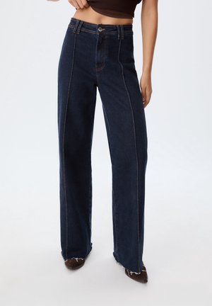 Frau trägt hoch taillierte, dunkelblaue Jeans mit weitem Bein und rohen Säumen sowie braune spitze Schuhe, zieht ein dunkles Oberteil leicht hoch.