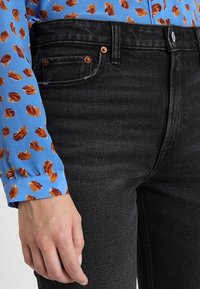 Jeans in denim nero con una vestibilità slim, design a cinque tasche, hardware in tono rame e una texture sottile. Abbinati a una camicetta blu con motivi marroni.