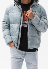 Aufgeblasene, hellblaue Jacke mit Reißverschluss und Kapuze, ausgestattet mit elastischen Bündchen und einer strukturierten Oberfläche, getragen über einem schwarzen Grafik-T-Shirt.