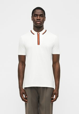 Άνδρας που στέκεται προς τα εμπρός, φοράει λευκό πόλο με κοντά μανίκια, γιακά με φερμουάρ με πορτοκαλί και μαύρες ρίγες και γκρι παντελόνι.