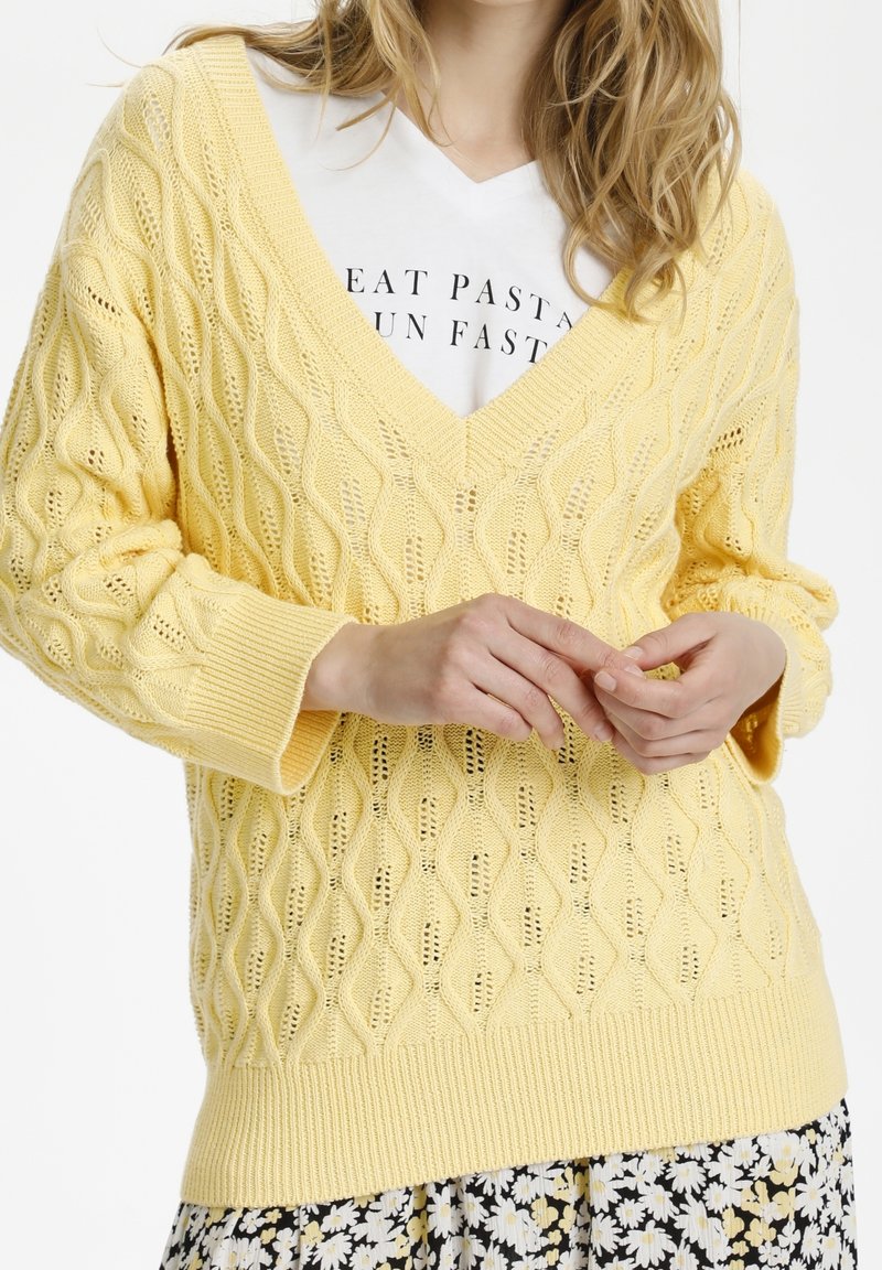 Helle gelbe Strickjacke mit V-Ausschnitt und strukturiertem Rautenmuster, mit gerippten Bündchen und Saum. Getragen über einem weißen T-Shirt mit Grafik.