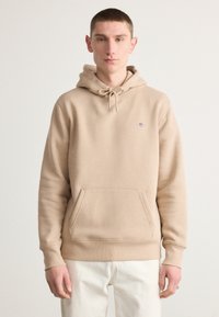 Beige hooded sweatshirt made of soft cotton blend fabric, featuring a kangaroo pocket and drawstring hood. Simple logo embroidered on chest.