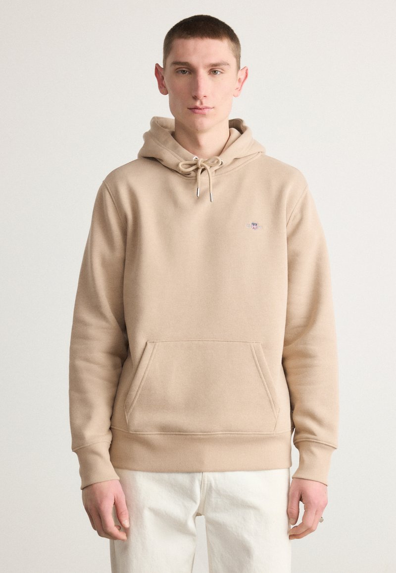 Beige hooded sweatshirt made of soft cotton blend fabric, featuring a kangaroo pocket and drawstring hood. Simple logo embroidered on chest.
