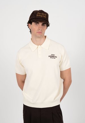 Mand iført en cremefarvet kortærmet polo med logoet "Prohibited Racing Club" og en brun kasket med teksten "The Art of Being Late."