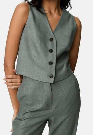 Ärmellose graue Weste mit Knöpfen, kombiniert mit passenden hochgeschnittenen Hosen, getragen von einer Person mit vor der Brust verschränkten Armen.