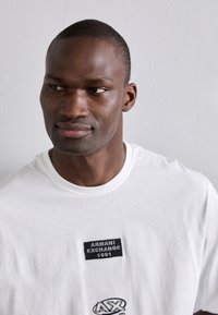 Fehér pamut T-shirt, fekete applikált címkével, rajta az "ARMANI EXCHANGE 1991" felirattal és egy kör alakú logóval az alsó elején, kerek nyakkivágással.