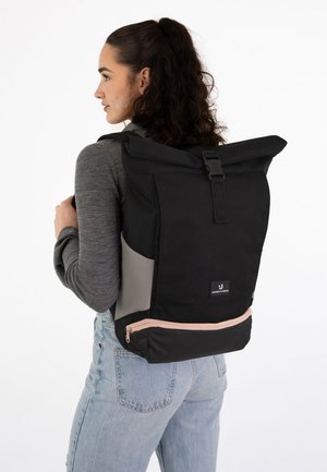 Mujer con cabello oscuro y rizado lleva un suéter gris y vaqueros azul claro, llevando una mochila negra grande con cierre enrollable y bolsillos laterales.