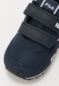 Tummansininen lenkkari, jossa on tarranauhat, teksturoitu mokkapinta, sileä materiaali ja valkoiset logoelementit. Kumikorko ja ompeluyksityiskohdat.