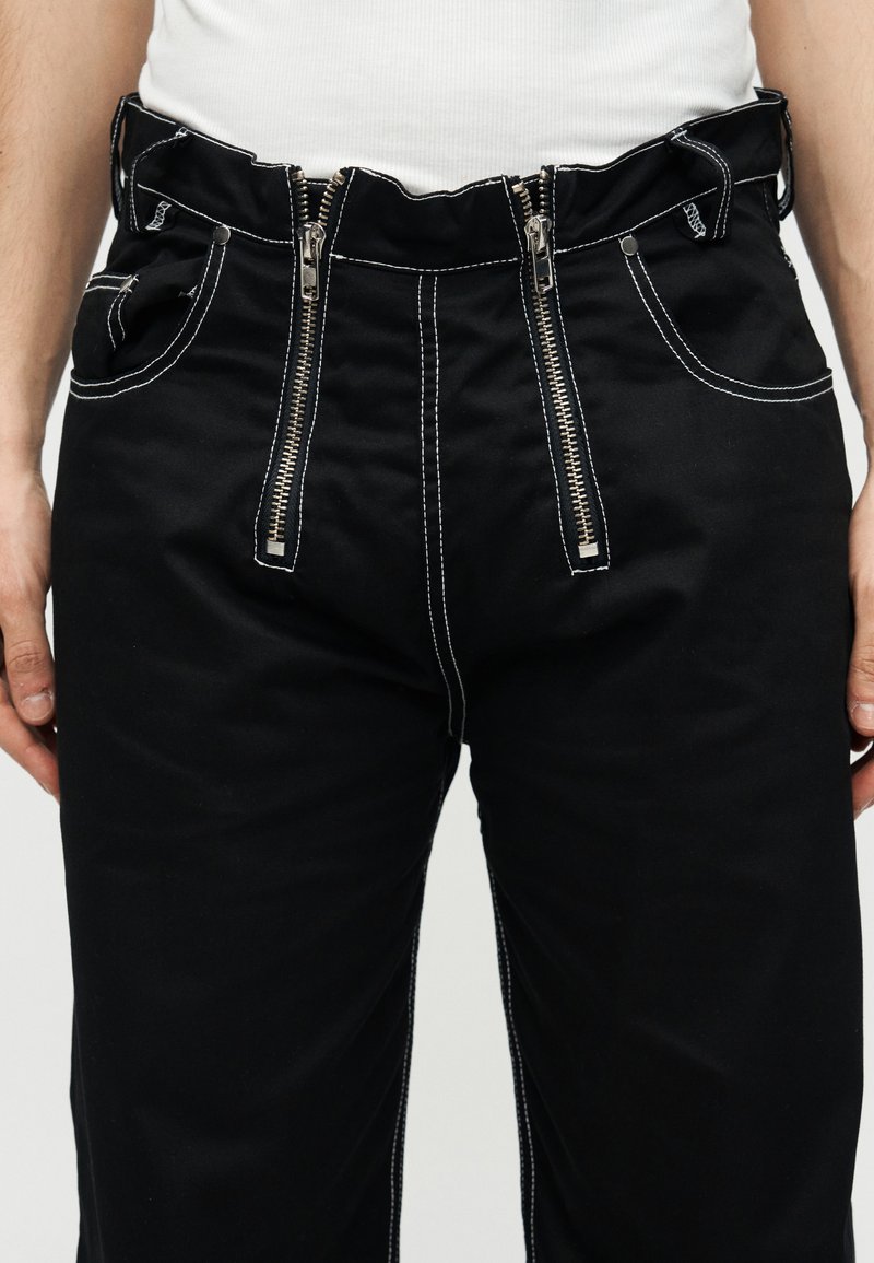 Black pants with two vertical front zippers and white contrast stitching worn with a white top, showing waist to mid-thigh area.
