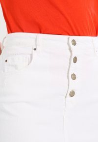 Gonna di jeans bianca a vita alta, caratterizzata da cinque bottoni in tono argento lungo la parte anteriore, e una superficie testurizzata con un leggero sfilacciamento ai bordi.