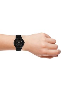 Montre analogique noire avec un cadran rond, bracelet en silicone noir texturé, marqueurs d'heure dorés et un design minimaliste au poignet.