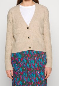 Beige cable-knit cardigan with a v-neck, featuring three large black buttons and ribbed hem, worn over a colorful patterned skirt.