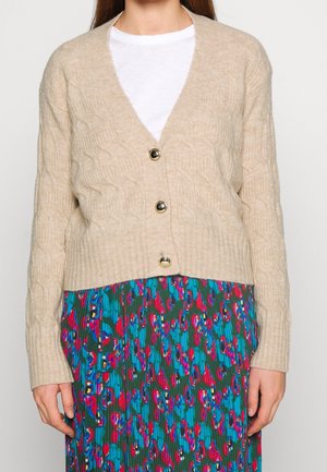 Beige kabelbrei cardigan met V-hals, voorzien van drie grote zwarte knopen en geribde zoom, gedragen over een kleurrijke patroonrok.