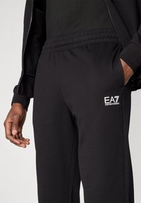 Μαύρο παντελόνι φόρμας με ελαστική μέση, πλαϊνές τσέπες και λευκό λογότυπο EA7. Μαλακό ύφασμα με ματ υφή, εφαρμοστή γραμμή.