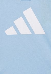 T-shirt azzurro chiaro a maniche corte con un grande logo bianco a tre strisce sulla parte anteriore. La texture del tessuto è liscia e morbida.