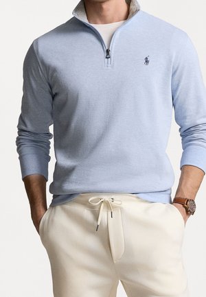 Mężczyzna w jasnoniebieskim swetrze z krótkim zamkiem i małym logo na piersi, kremowych spodniach ze sznurkiem oraz zegarku na nadgarstku, z rękami w kieszeniach.