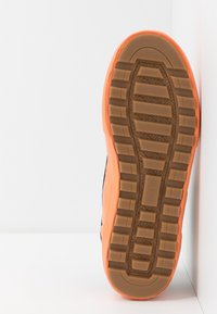 Die Außensohle des Sneakers weist ein strukturiertes braunes Gummimuster mit einem orangefarbenen Akzent auf. Das Design umfasst eine glatte Kante und dezente Markenkennzeichnung.