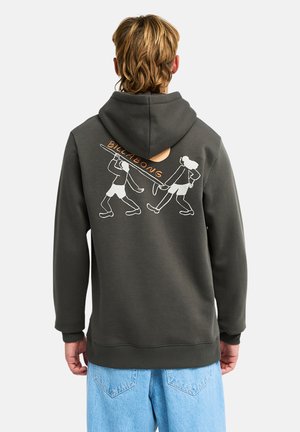 Persona che indossa una felpa scura con cappuccio con una grafica di surf e il testo "Billabong" sulla schiena, abbinata a jeans azzurri, di spalle.
