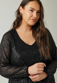 Zwarte sequined top met een V-halslijn, lange mouwen en een textuurstof. Heeft een losse pasvorm en gerimpelde manchetten aan de mouwen.