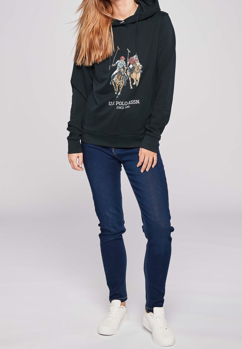 Sweat à capuche noir avec un motif représentant des cavaliers, associé à un jean bleu foncé et des baskets blanches. Tenue décontractée pour le quotidien.