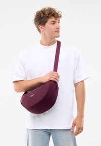 Burgundy axelväska med en mjuk yta, halvmåneform, justerbar rem och varumärkesdetaljer, hålls av en person i en vit T-shirt.