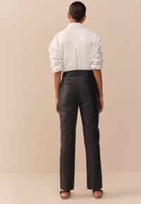 Pantalon en cuir noir à coupe droite, doté de deux poches arrière et d'une texture lisse, assorti à une chemise blanche.