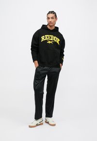 Čierna mikina s kapucňou a žltým logom "Reebok", v kombinácii s čiernymi športovými nohavicami a krémovými teniskami s gumovou podrážkou a čiernymi akcentmi.